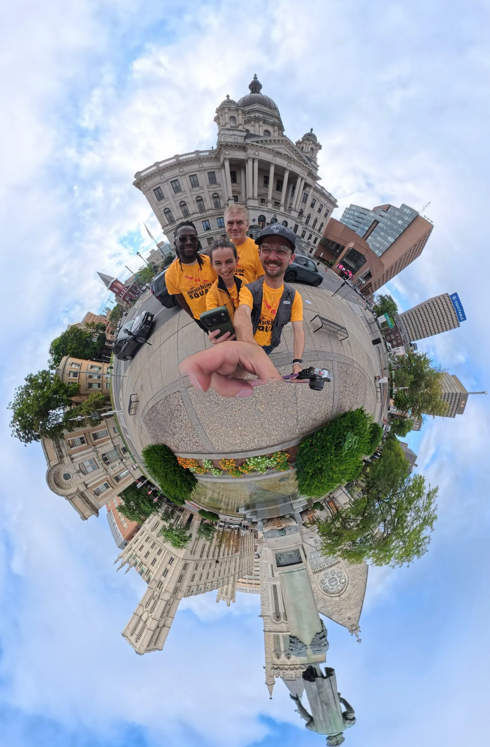 A 360 degree tiny-planet photo of the team in yellow shirts standing in the circle, with the surrounding buildings wrapped around them