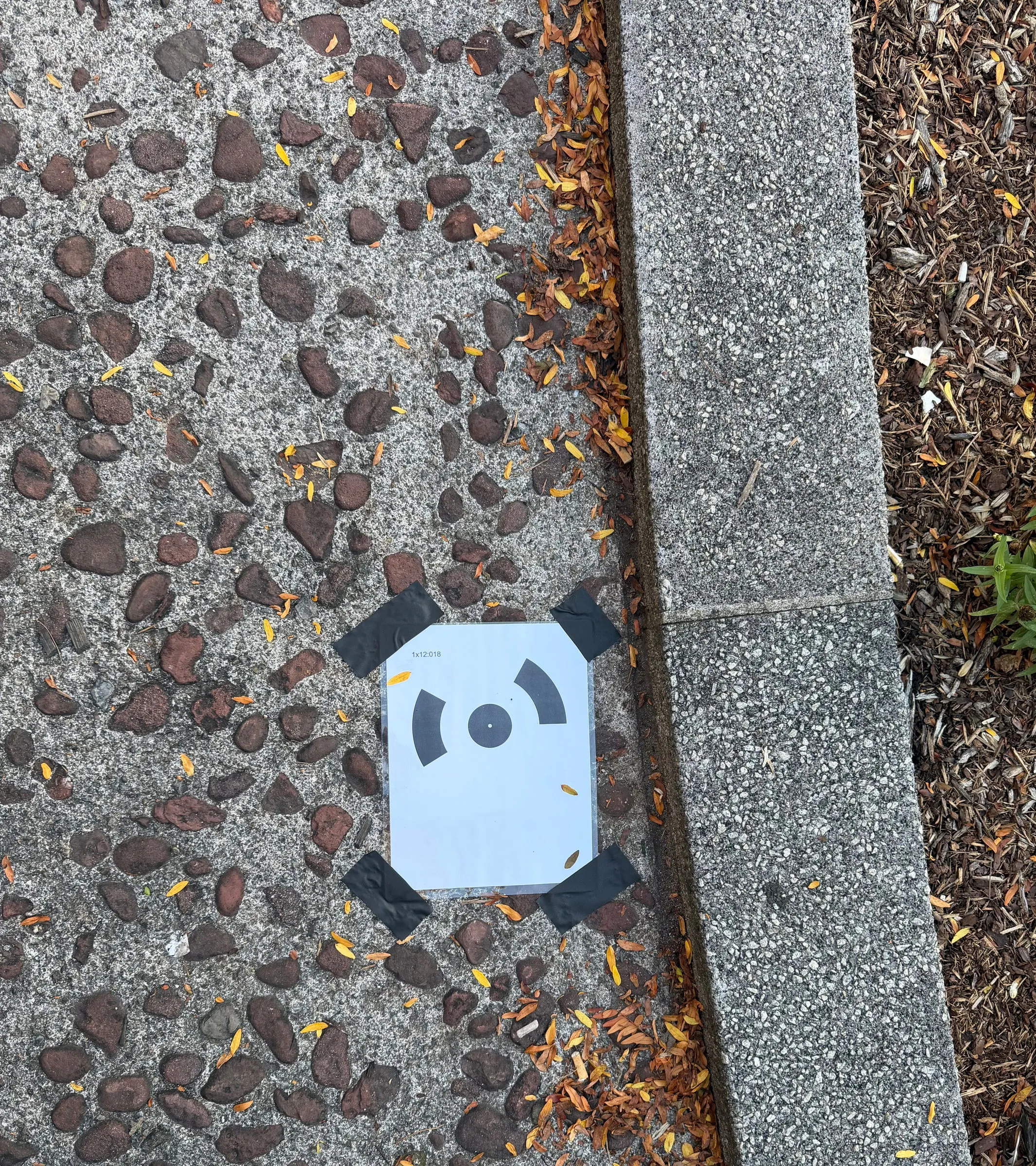 A square black and white scanning marker taped to the pebbled paving at the edge of a curb