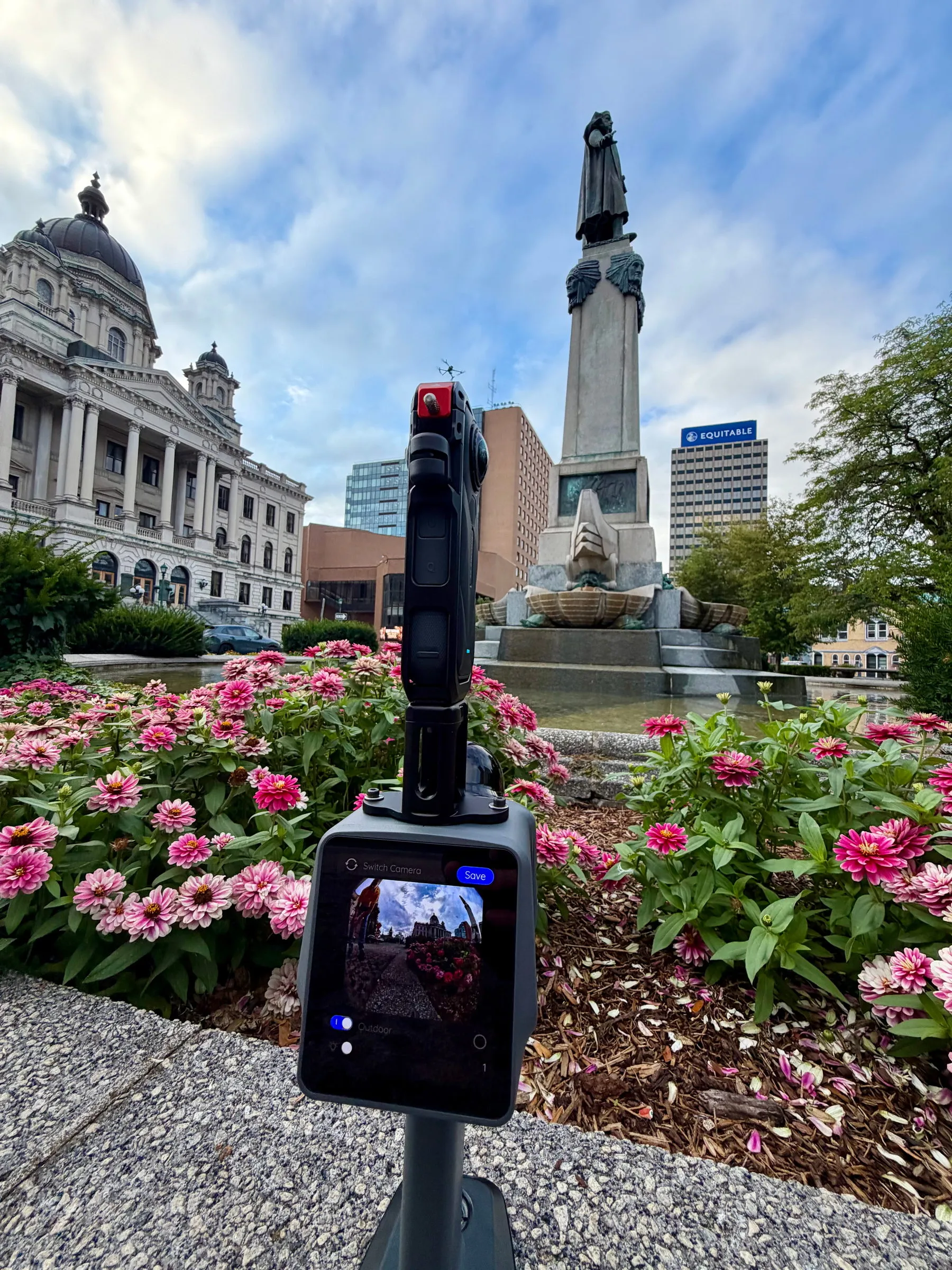 A capture rig on a pole in a bed of pink flowers, facing the monument and courthouse
