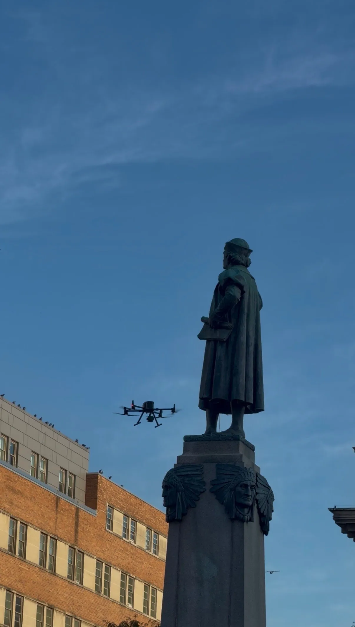 The monument against a blue sky with a brick building behind and the drone beside the figure
