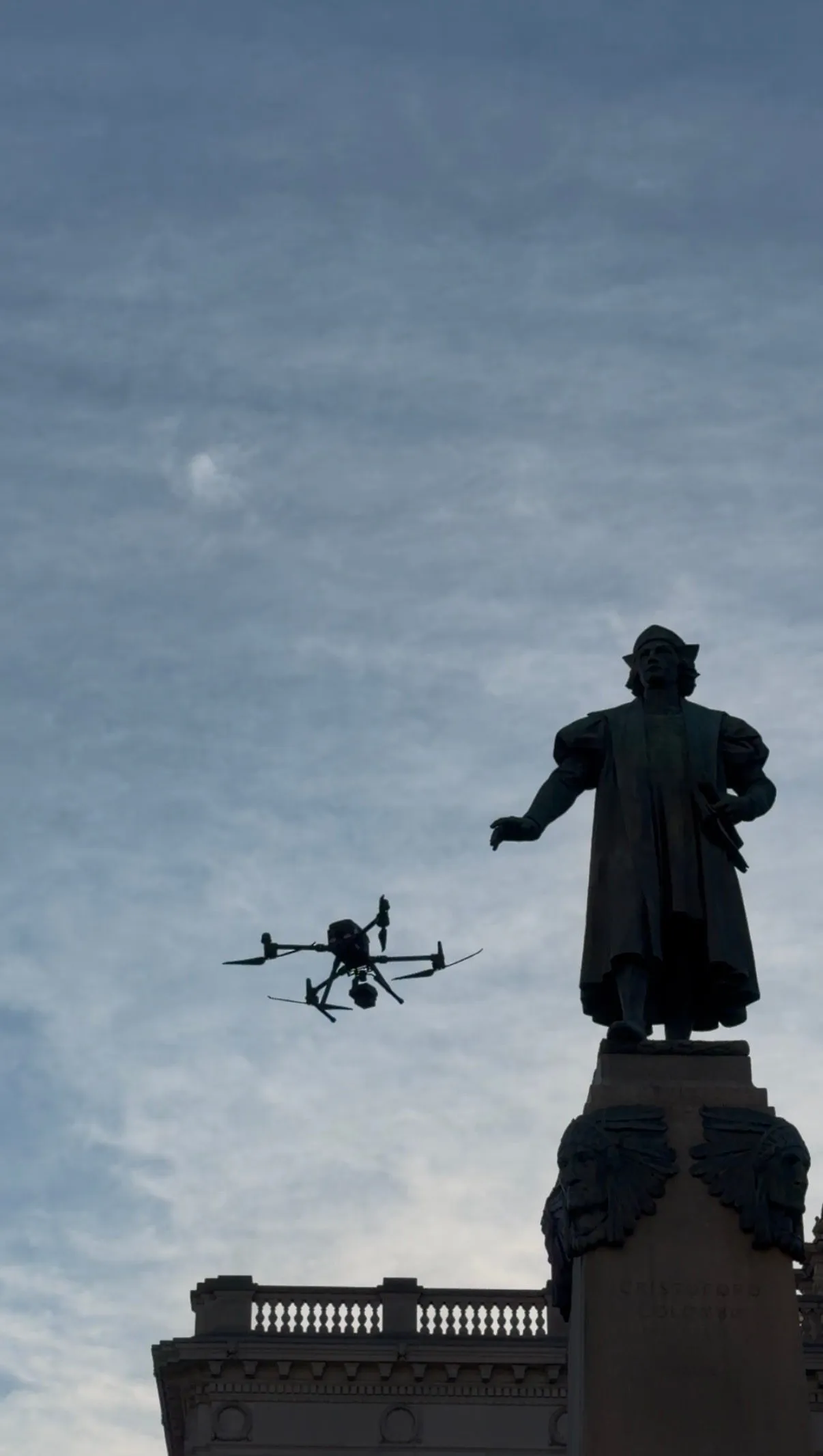Silhouette of the bronze figure against an overcast sky, with the drone hovering beside it