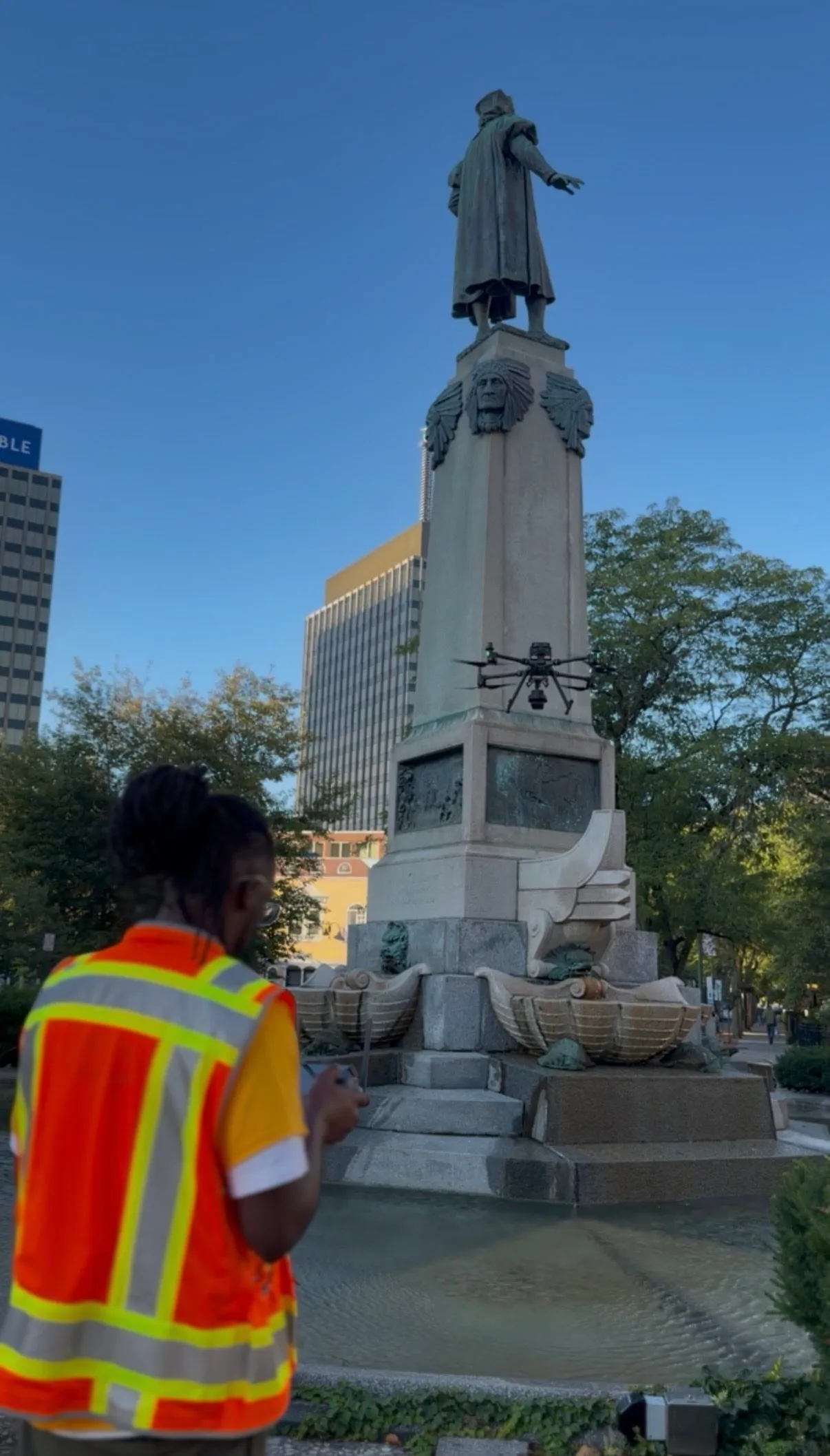 A team member in a high-visibility vest flying the drone at the base of the monument