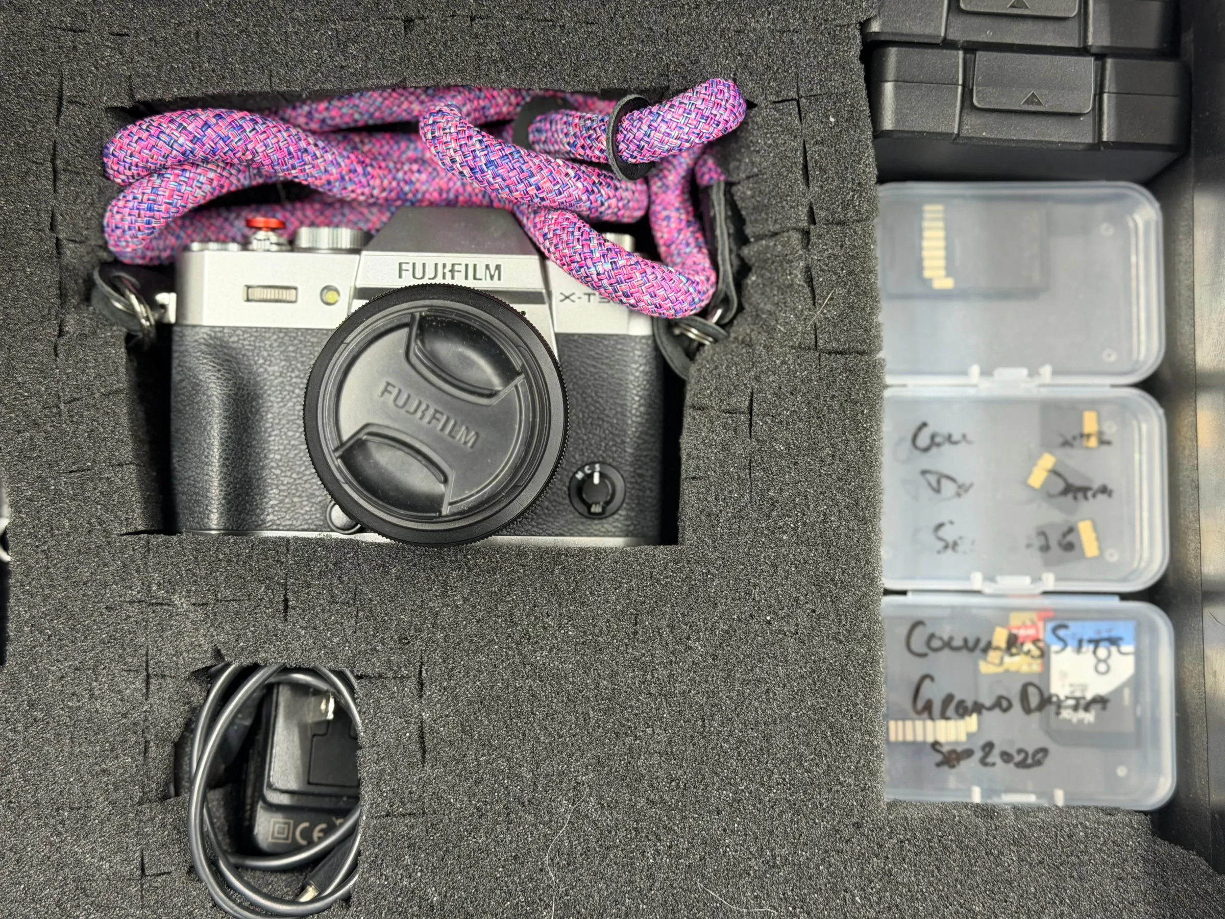 A camera with a pink strap in a foam-lined case beside small labeled boxes of memory cards