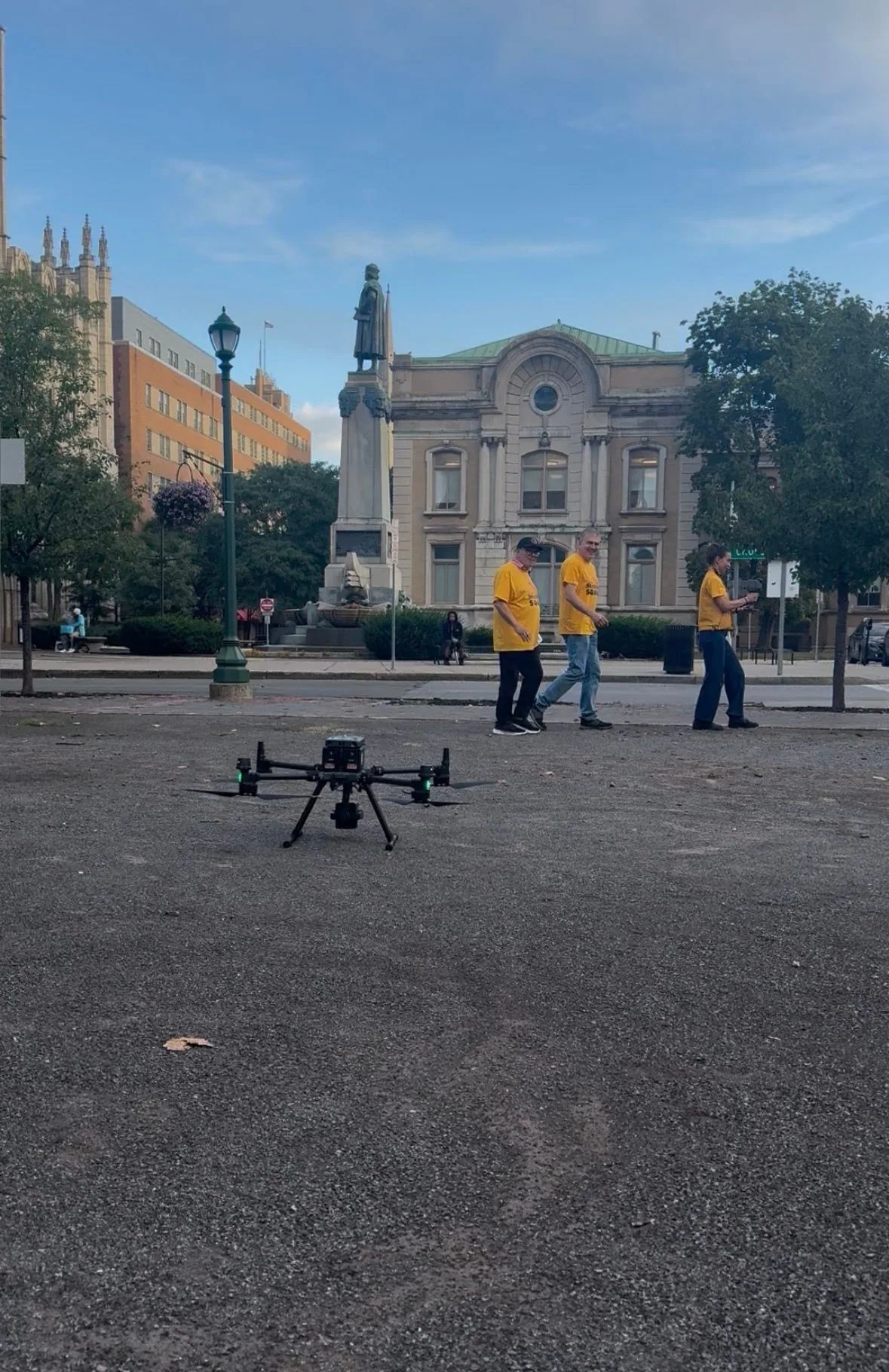 The drone on the gravel of the Powelson Site with three team members walking behind it, and the monument and fountain across the street